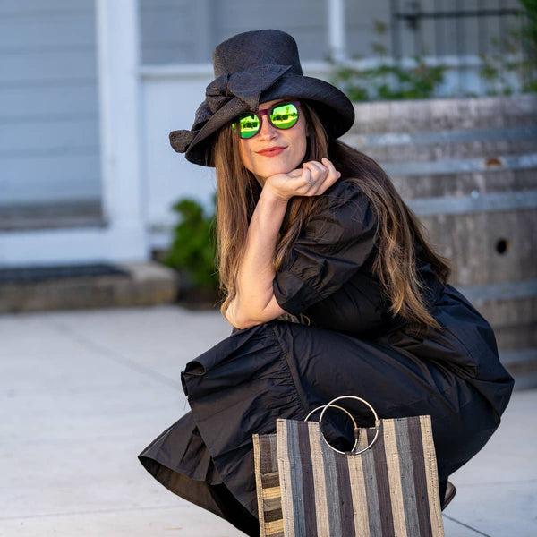 Woman in black outfit with sunglasses and hat, holding a striped bag on a sidewalk.
