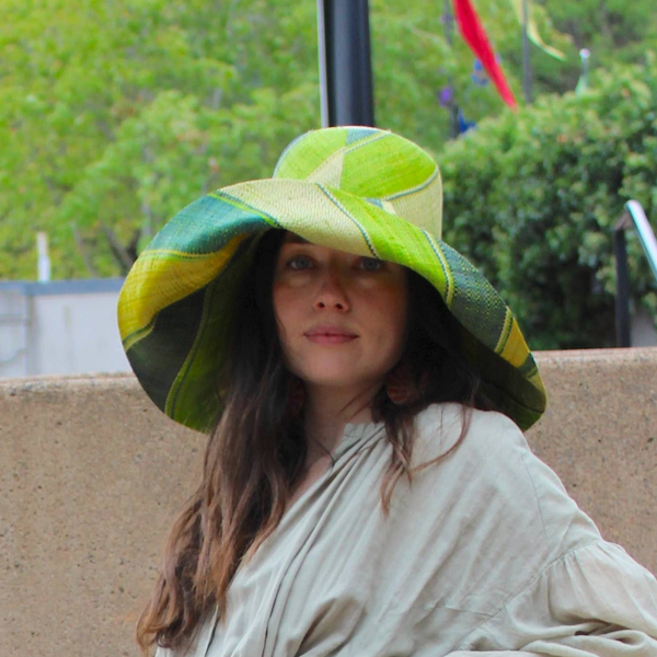Woman in a long light green dress and large hat sitting on stone steps outdoors.