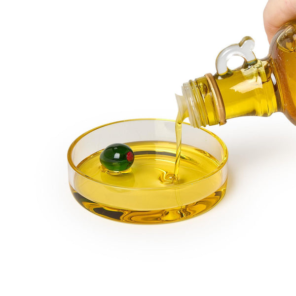Olive oil being poured from a bottle into a glass bowl with an olive on a white background