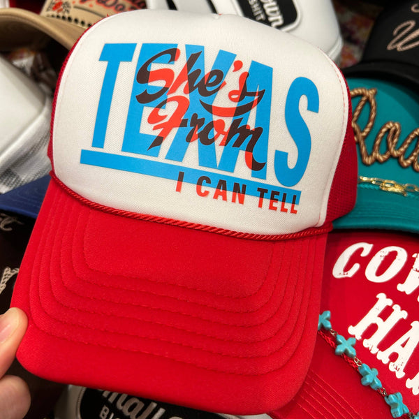 Red and white cap with 'She's from Texas I can tell' text, surrounded by other caps.